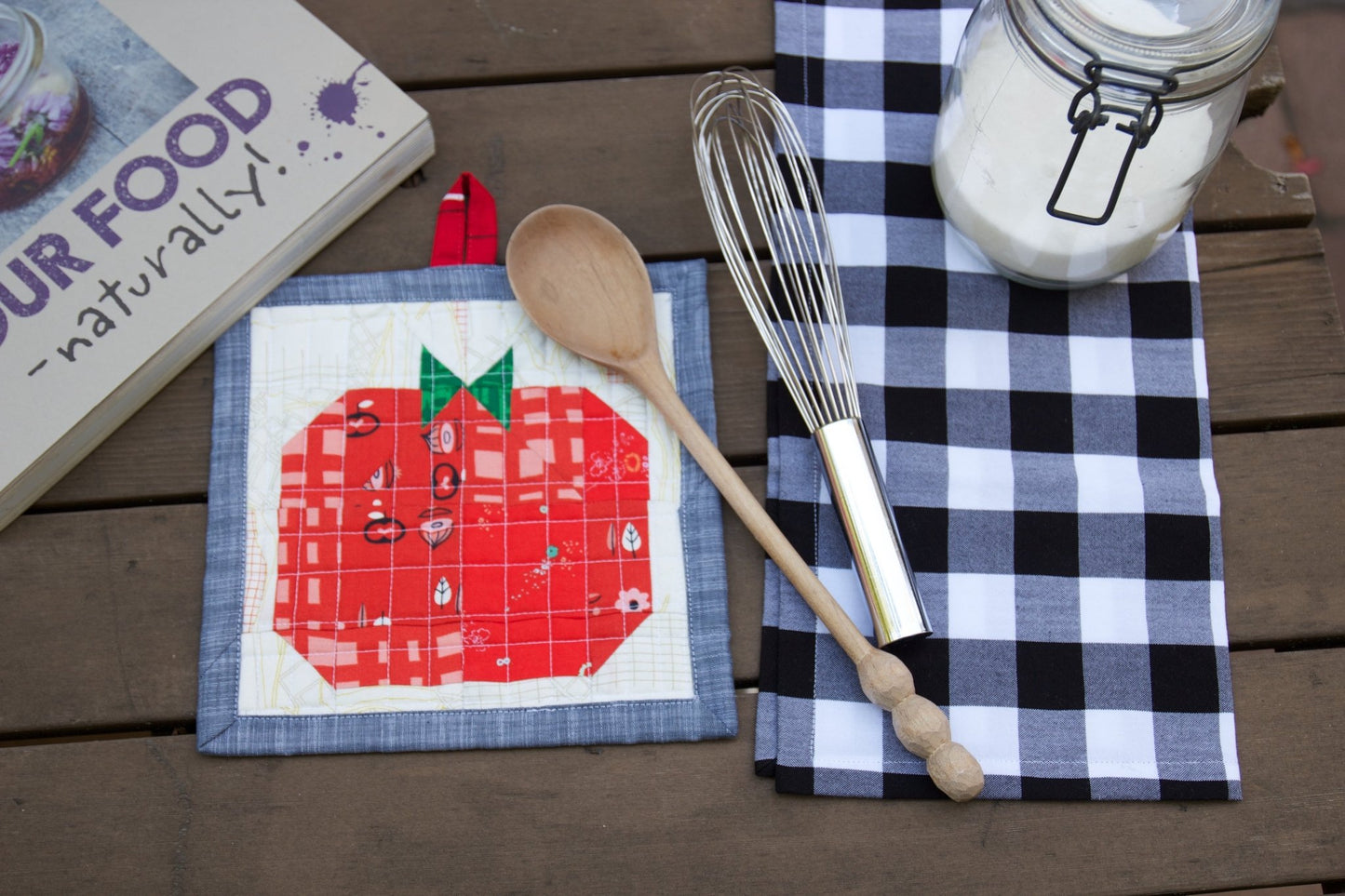 Quilted tomato pot holder on a table with cloth napkin and utensils.