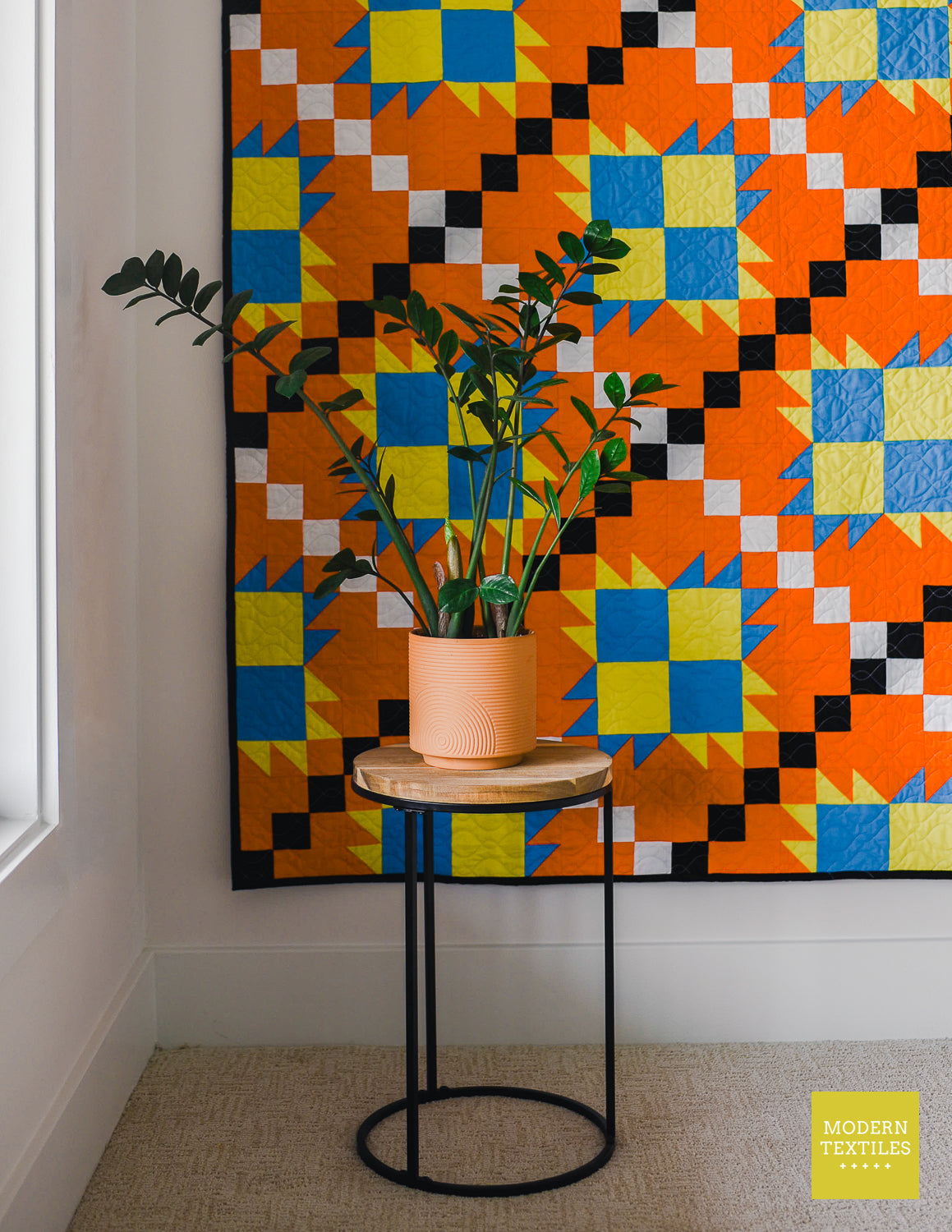 Orange, black, white, blue and lime green patchwork quilt hanging on white wall with plant on table in front of the quilt. Modern Textiles branding on the lower right hand corner.