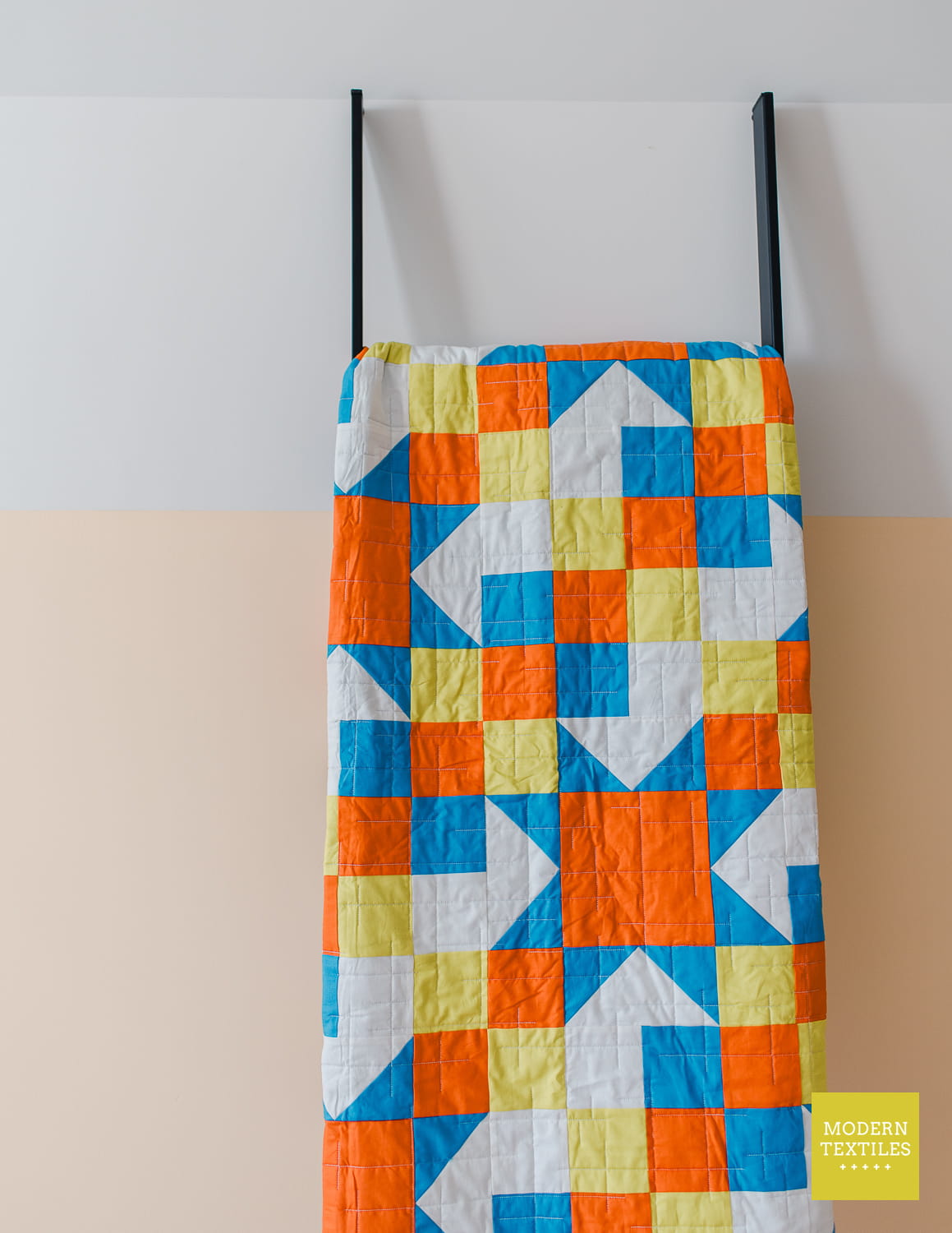 A colorful quilted textile hanging from black metal ladder against a two-tone wall, featuring a geometric pattern of orange, blue, yellow, and white blocks.