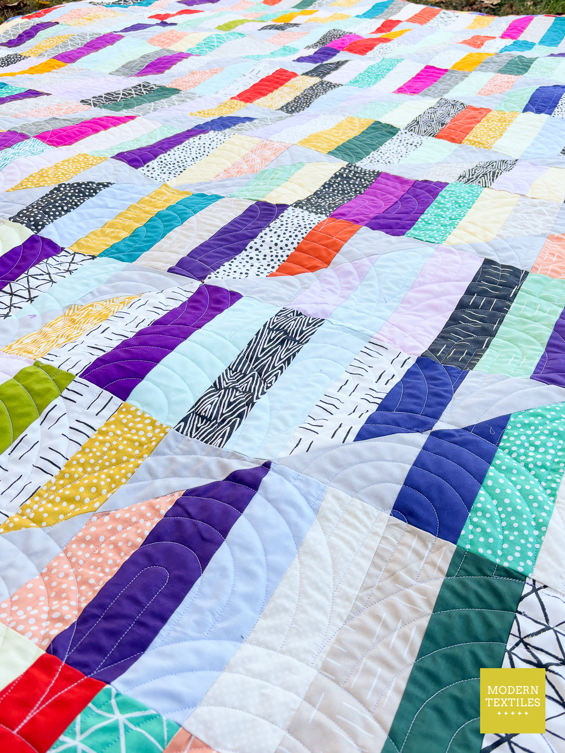 A colorful patchwork quilt laid out flat, featuring a variety of vibrant and pastel patterned fabric blocks. The quilt fills the entire frame, with a yellow 'MODERN TEXTILES' logo in the bottom right corner.
