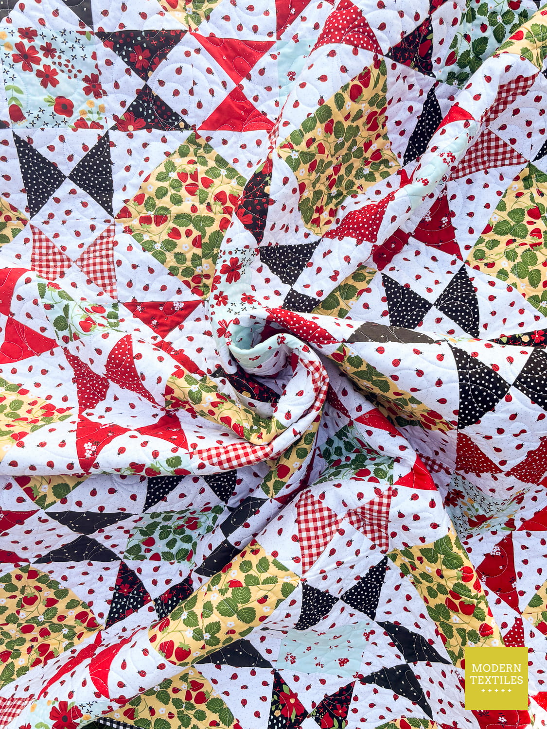 A close-up of the quilt featuring a festive summery fabric design in red, green, black, and white patterns including gingham, polka dots, floral prints, and strawberries, styled with a central swirl. 'Modern Textiles' logo in lower right hand corner.