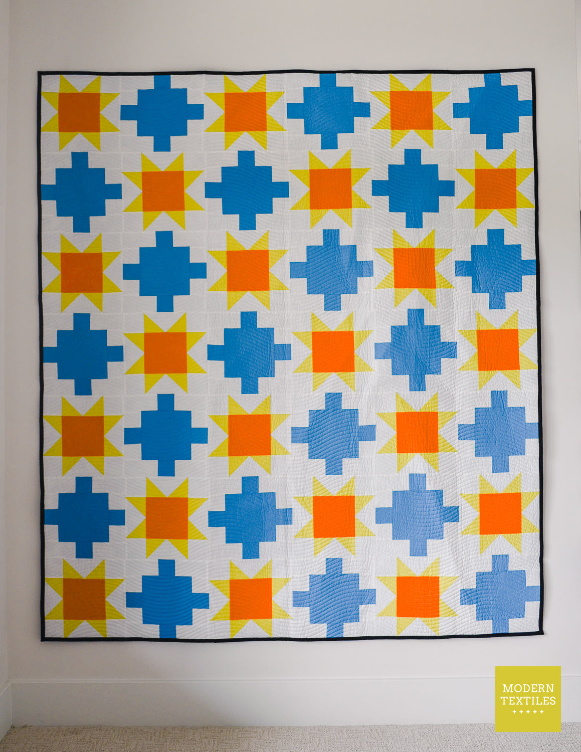 Blue patchwork quilt with sawtooth stars in orange and yellow-green with white background quilt on a white wall.