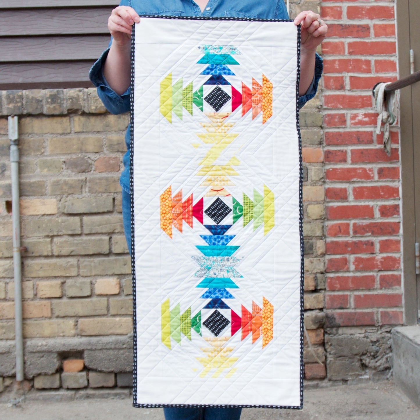 Pineapple Color Wheel quilt table runner held up against a brick wall, featuring bright rainbow pineapple motifs.