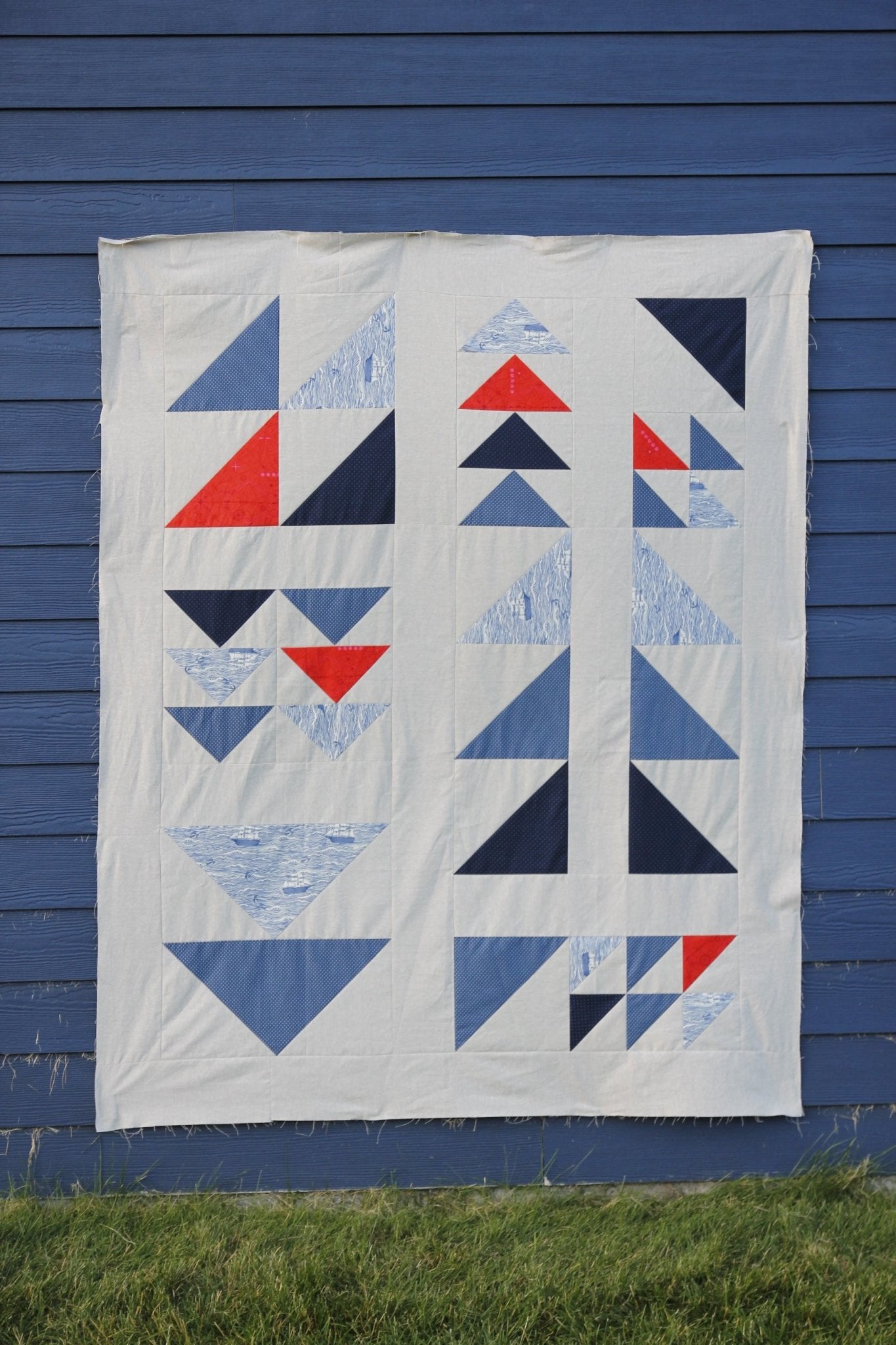 Finished Exploratory Quilt hanging outdoors against a blue wall, showing flying geese and half-square triangle blocks.