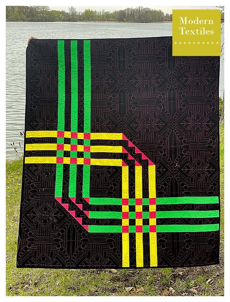 Pipeline Quilt laid flat on grass near a lake, showcasing bright green, yellow, and red strip accents on a dark background.