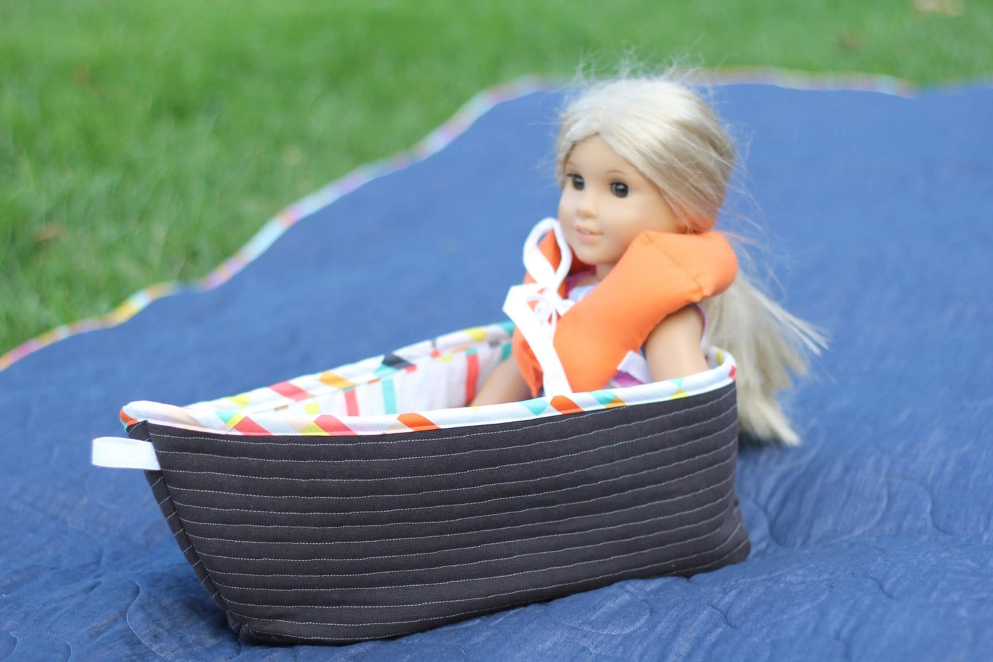 Toy doll sitting inside quilted dinghy with patterned interior and orange life jacket.