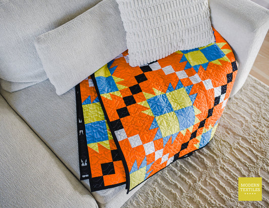 Orange, black, white, blue and lime green patchwork quilt  folded and diagonally on tan/grey couch with textured pillow on top of quilt and light grey textured rug on ground. Modern Textiles branding on the lower right hand corner.