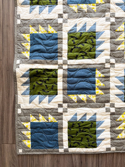 Corner of modern quilt showcasing geometric pattern featuring blue, green, and gray colors on a wooden surface.