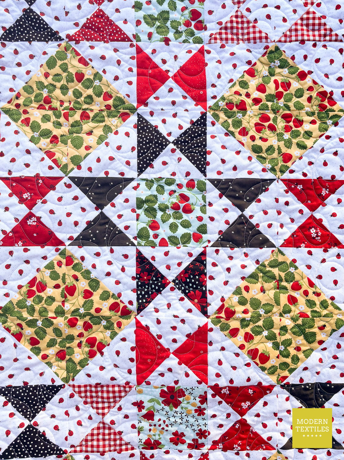 A close-up of the Ohio Star Block with red, black, green, and white patterned fabric, including strawberry prints, gingham, polka dots, and floral motifs.