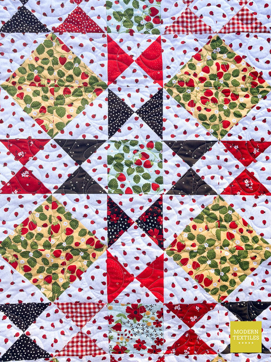 A close-up of the Ohio Star Block with red, black, green, and white patterned fabric, including strawberry prints, gingham, polka dots, and floral motifs.