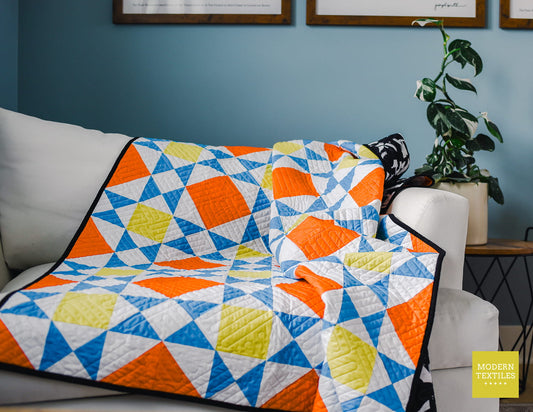 A colorful geometric quilt draped over a white sofa in a living room setting, featuring a pattern of orange, blue, yellow, and white triangles with black borders. A potted plant and framed wall art are visible in the background.
