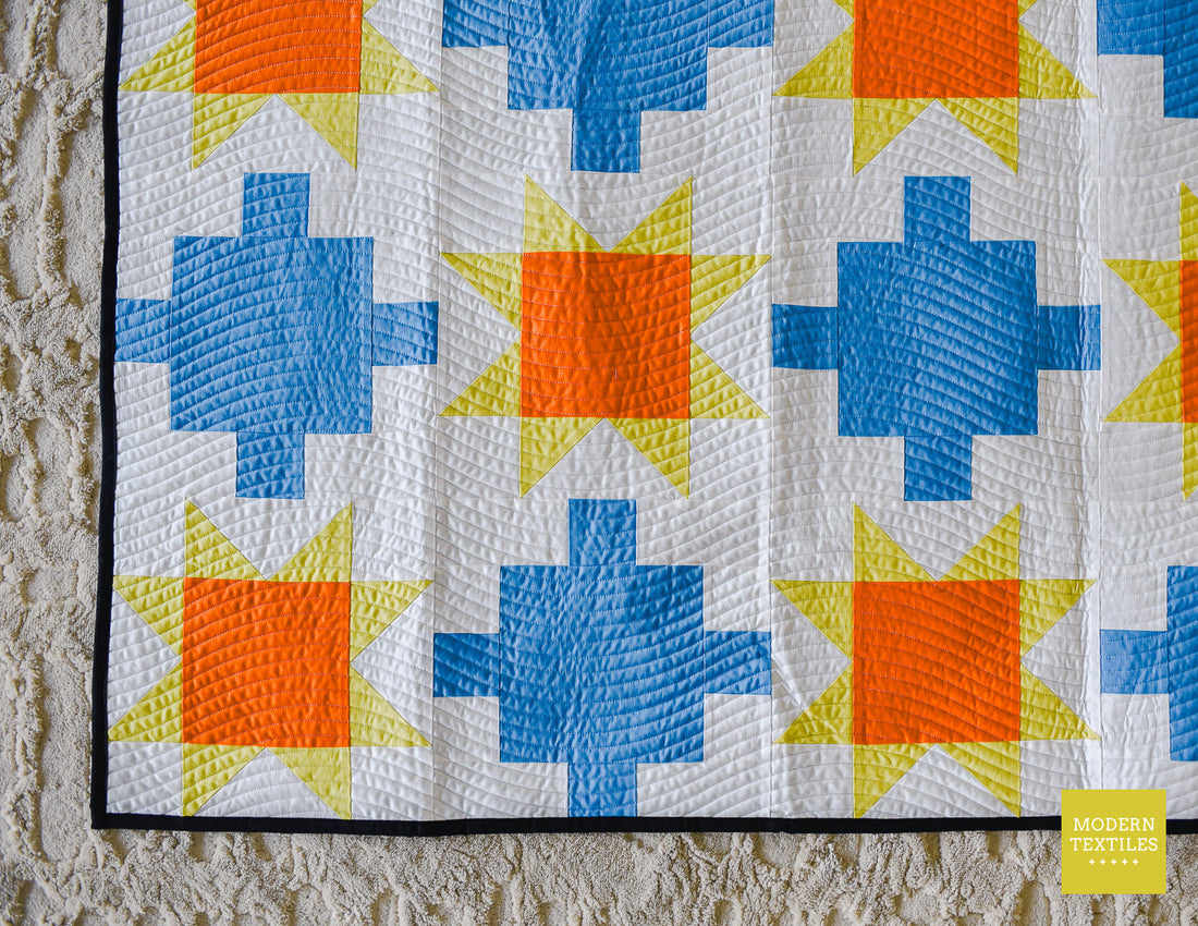 Close-up of blue patchwork quilt with sawtooth stars in orange and yellow-green with white background fabric and black binding fabric on top of neutral textured carpet.