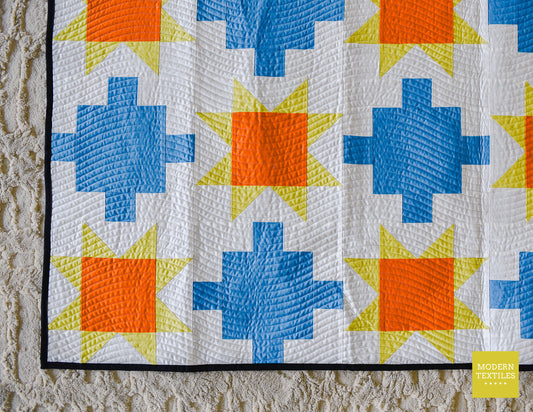 Close-up of blue patchwork quilt with sawtooth stars in orange and yellow-green with white background fabric and black binding fabric on top of neutral textured carpet.