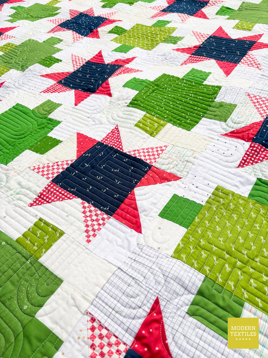An angled close up of the stars and lantern quilt to show off fun prints that are from fat quarters for the entire quilt top in navy, green, hot pink and a variety of whites and creams for teh background. Modern Textiles Logo in lower right corner.