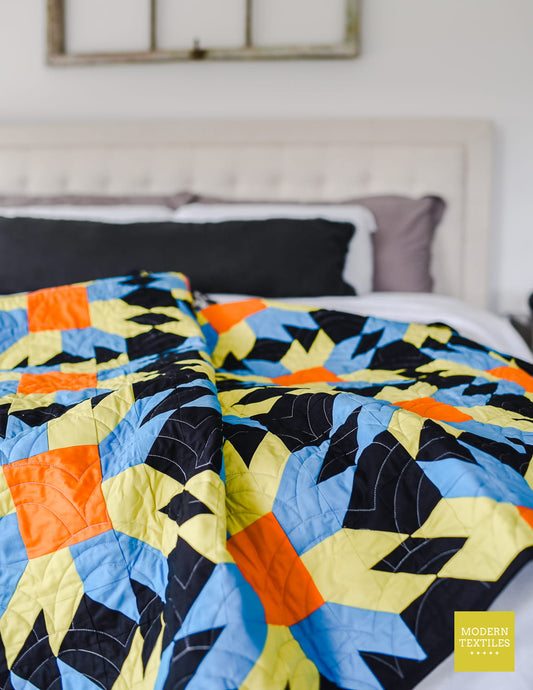 A colorful geometric quilt with blue, yellow, orange, and black patchwork draped over a bed with neutral headboard in background.