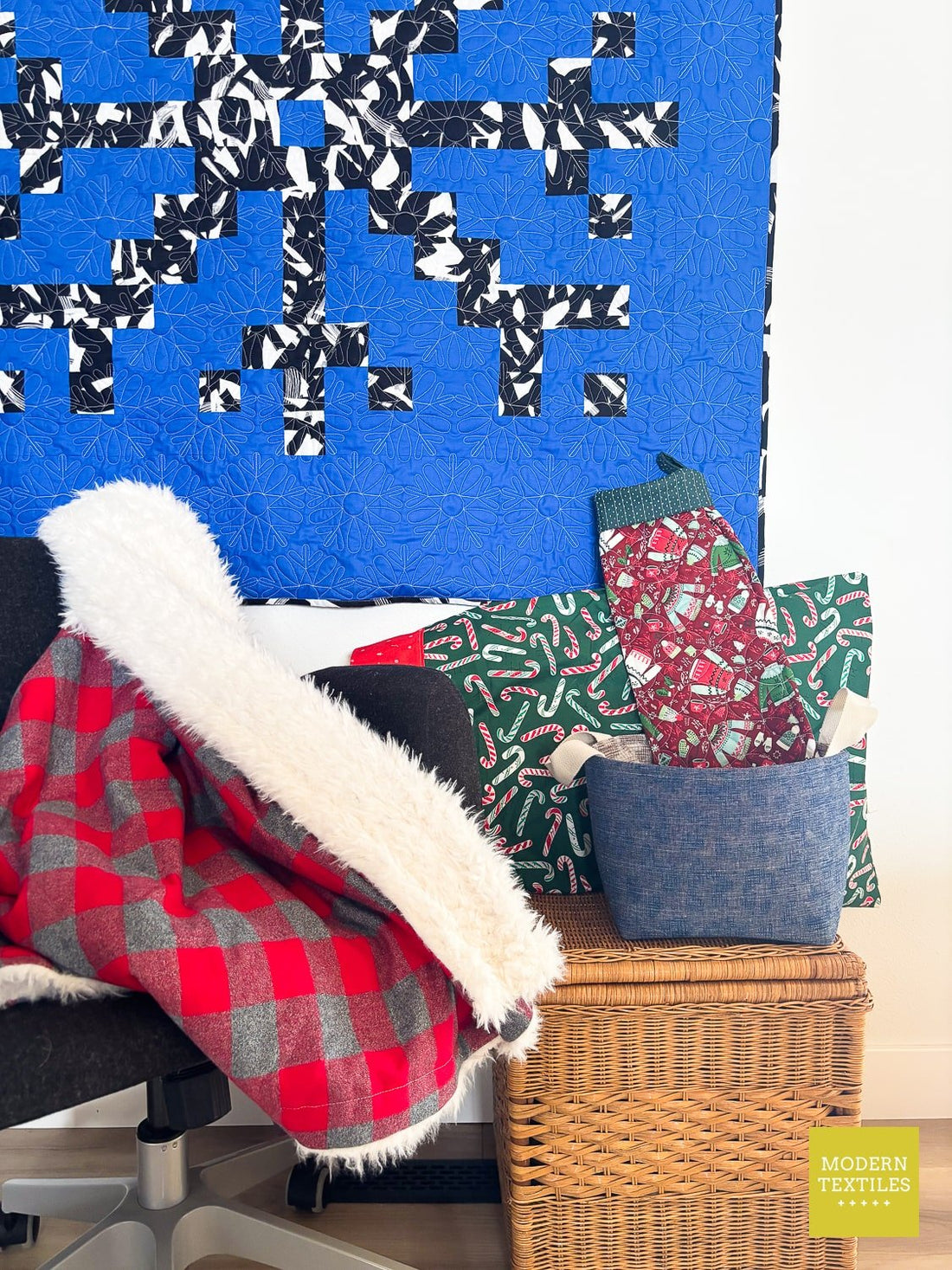 Holiday sewing display with a blue snowflake quilt, cozy red buffalo-plaid blanket, festive stocking, candy cane pillowcase and fabric storage basket.