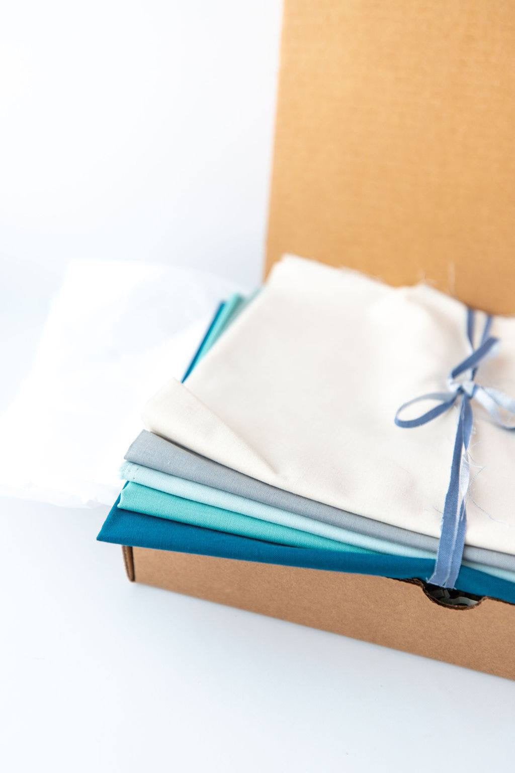 Fabric yardage in a folded bundle with white, grey, light blue, teal and turquoise wrapped with string and set onto an open box showcasing as a quilt kit.