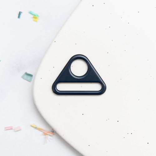 Navy blue triangular d-ring hardware on a white speckled surface with colorful items in the corner.