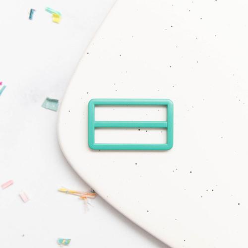 Turquoise rectangular tri glide buckle on a white speckled surface with colorful items in the background