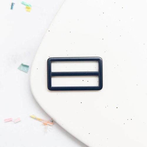 Navy blue rectangular tri glide buckle hardware on a white surface with colorful items in the background