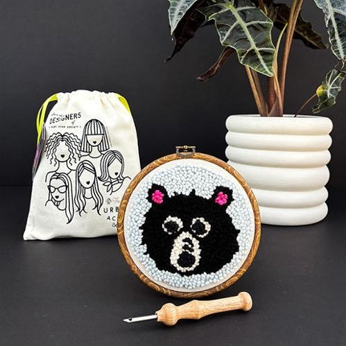 Embroidery hoop with a bear design, a drawstring bag, and a plant on a dark surface.