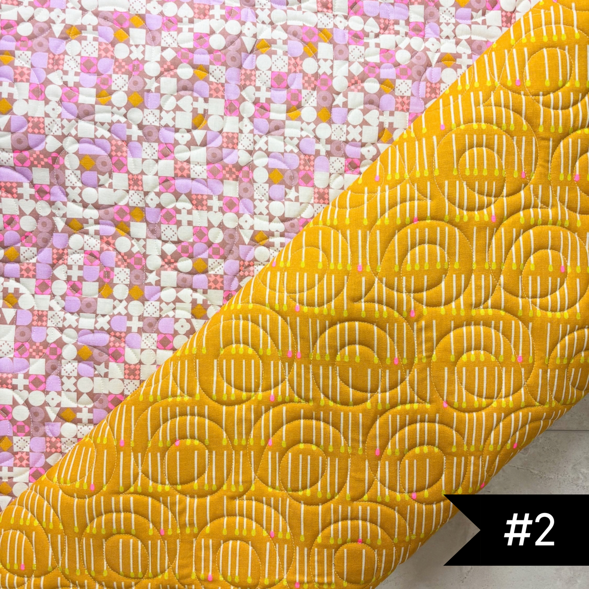 A quilted fabric panel shown diagonally, featuring a bright yellow lining fabric with white stitched soho quilt patnto and a pink, white, and yellow patchwork printed exterior. A black label in the bottom right corner reads '#2'.
