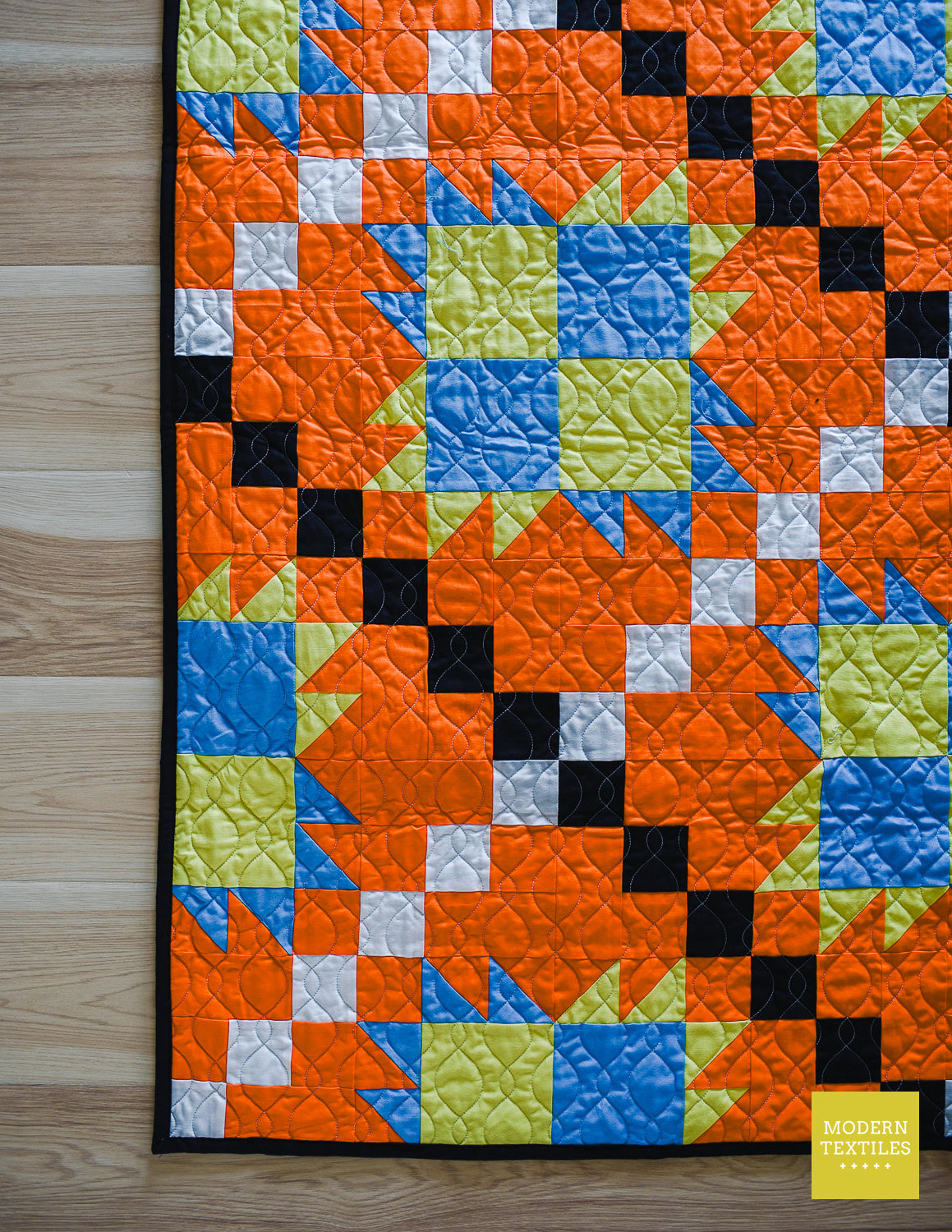 Close-up of orange, black, white, blue and lime green patchwork quilt crinkled on wood flooring, quilting texture is featured along with black binding on the bottom left hand corner . Modern Textiles branding on the lower right hand corner.