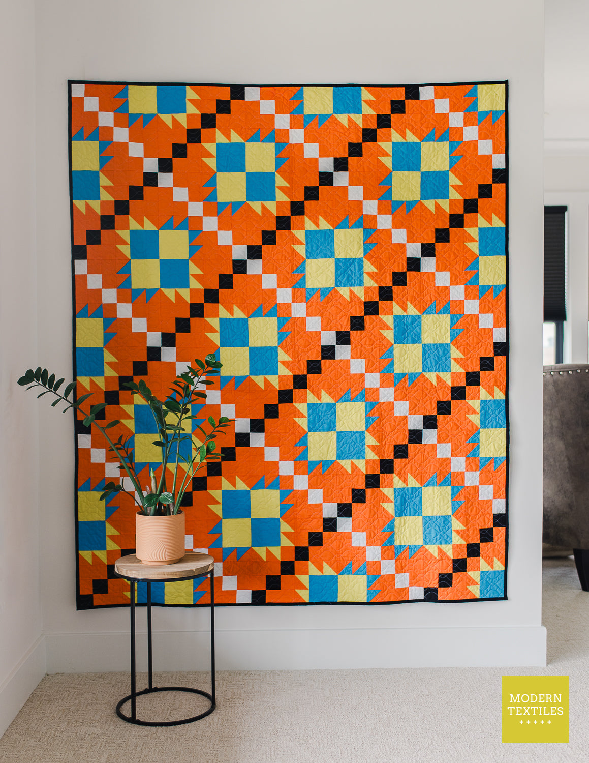 Orange, black, white, blue and lime green patchwork quilt hanging on white wall with plant on table in front of the quilt off to the lower left. Modern Textiles branding on the lower right hand corner.
