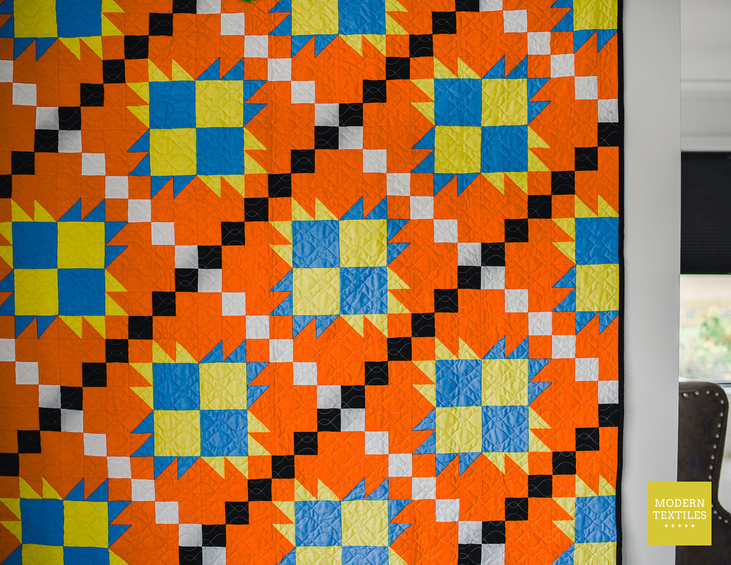 Close-up of orange, black, white, blue and lime green patchwork quilt hanging on white wall with chair and window in background. Modern Textiles branding on the lower right hand corner.