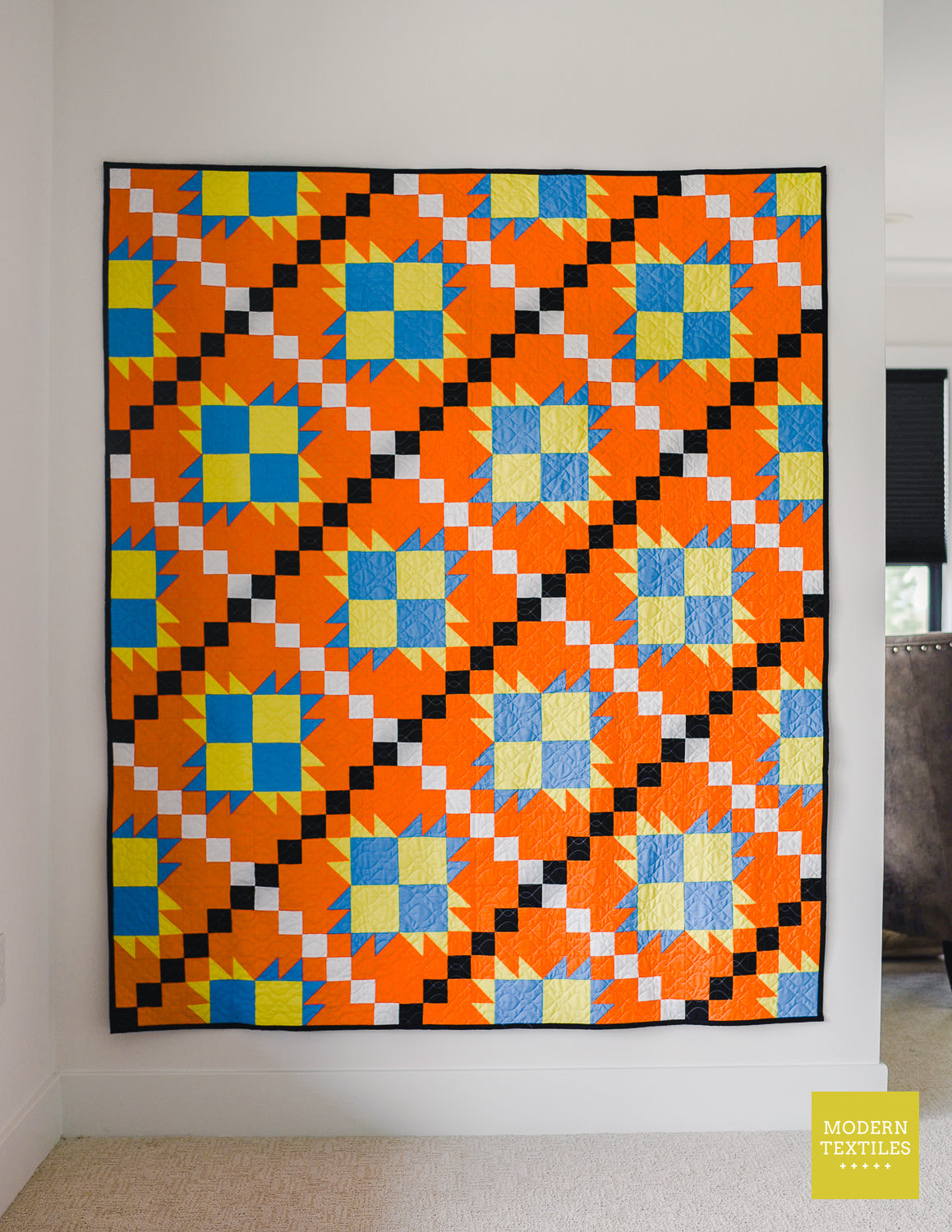 Orange, black, white, blue and lime green patchwork quilt hanging on white wall. Modern Textiles branding on the lower right hand corner.