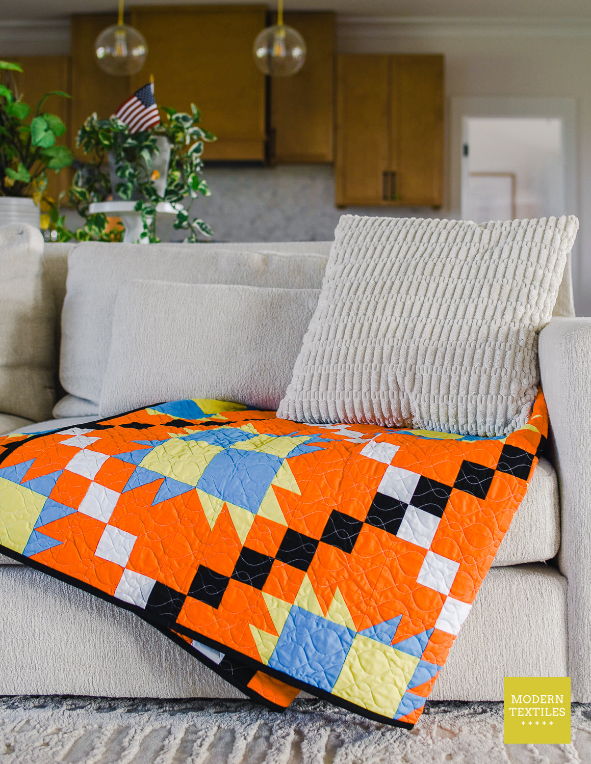 Orange, black, white, blue and lime green patchwork quilt  folded and diagonally on tan/grey couch with textured pillow on top of quilt and light grey textured rug on ground and kitchen cabinets, plants in background. Modern Textiles branding on the lower right hand corner.