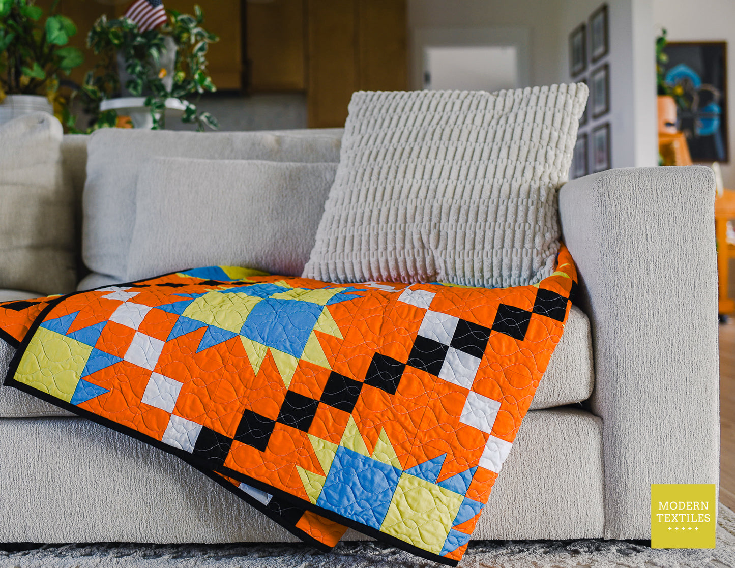 Orange, black, white, blue and lime green patchwork quilt  folded and diagonally on tan/grey couch with textured pillow on top of quilt and light grey textured rug on ground and kitchen cabinets, plants in background. Modern Textiles branding on the lower right hand corner.