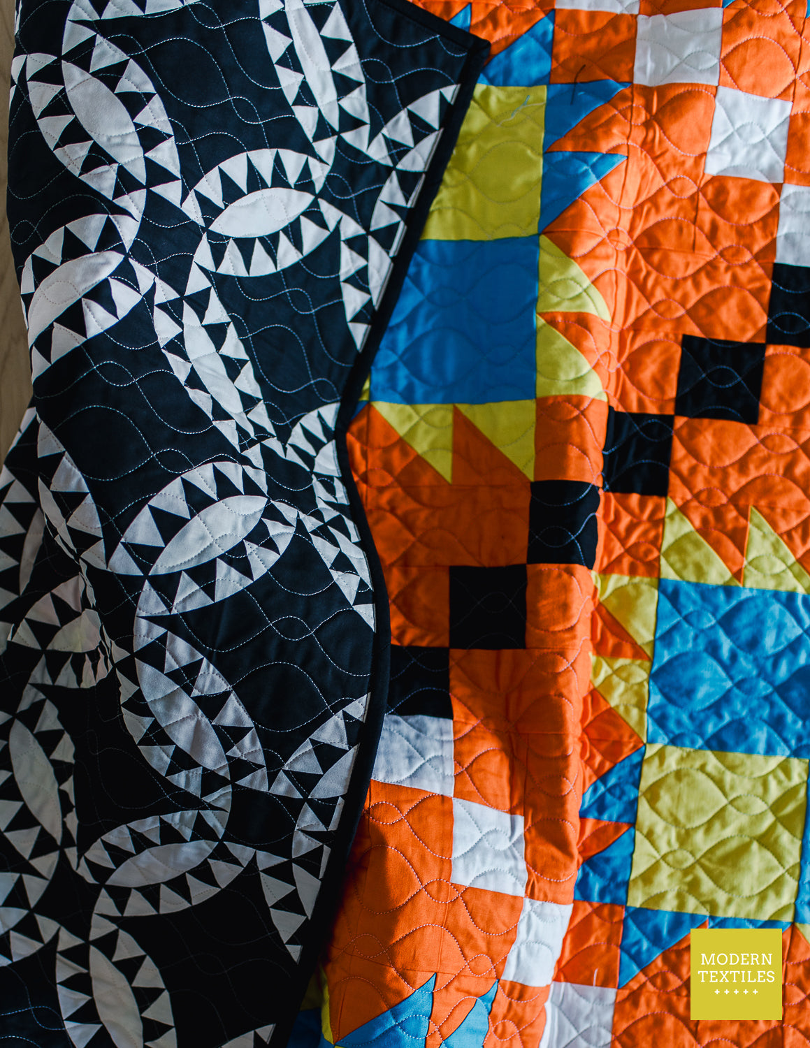Orange, black, white, blue and lime green patchwork quilt crinkled on ground with left hand side of quilt folded over to showcase black and white geometric pickle-dish backing fabric and looping quilting texture. Modern Textiles branding on the lower right hand corner.