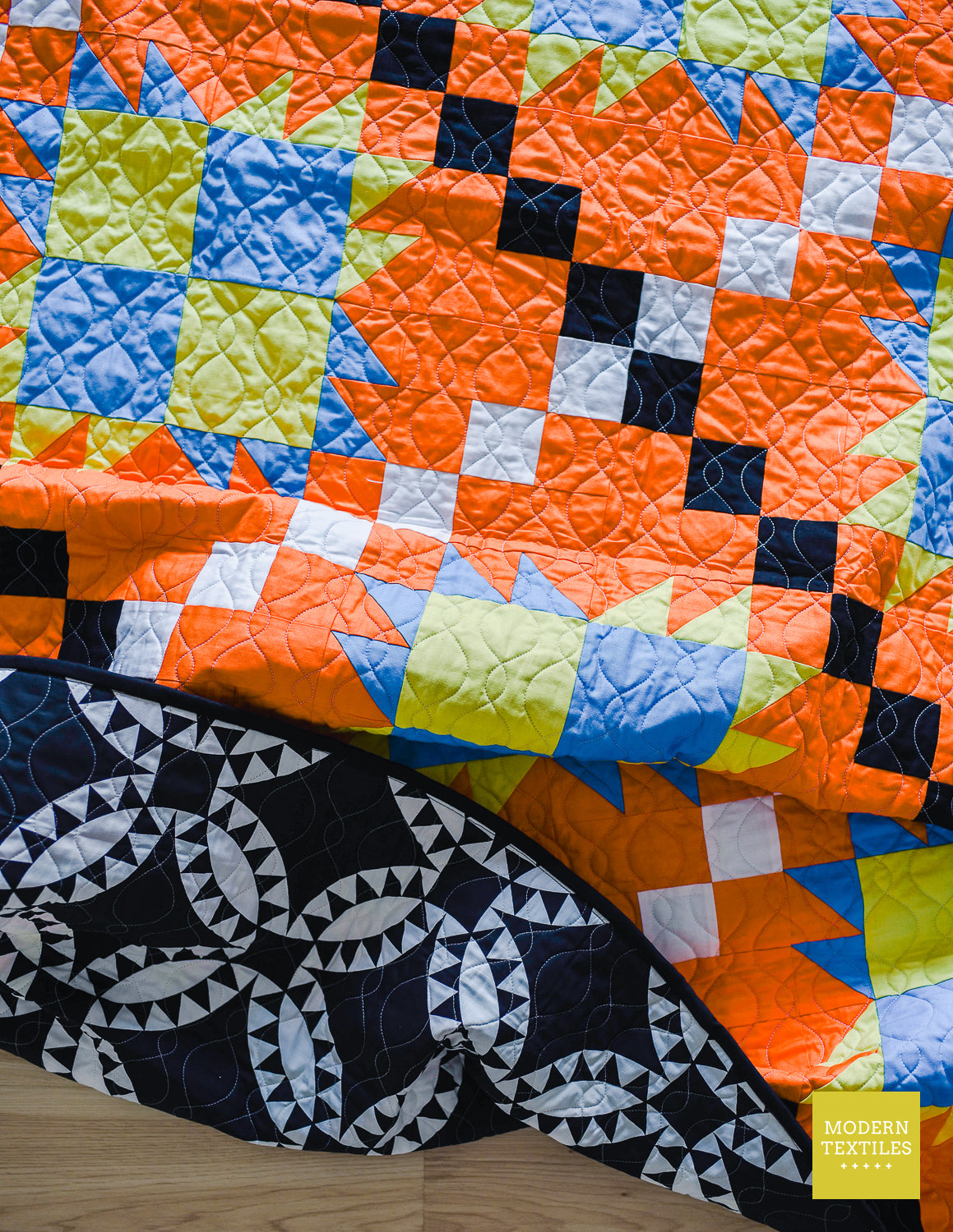 Orange, black, white, blue and lime green patchwork quilt crinkled on ground with left hand side of quilt folded over to showcase black and white geometric pickle-dish backing fabric and looping quilting texture with wood floor showing underneath quilt. Modern Textiles branding on the lower right hand corner.