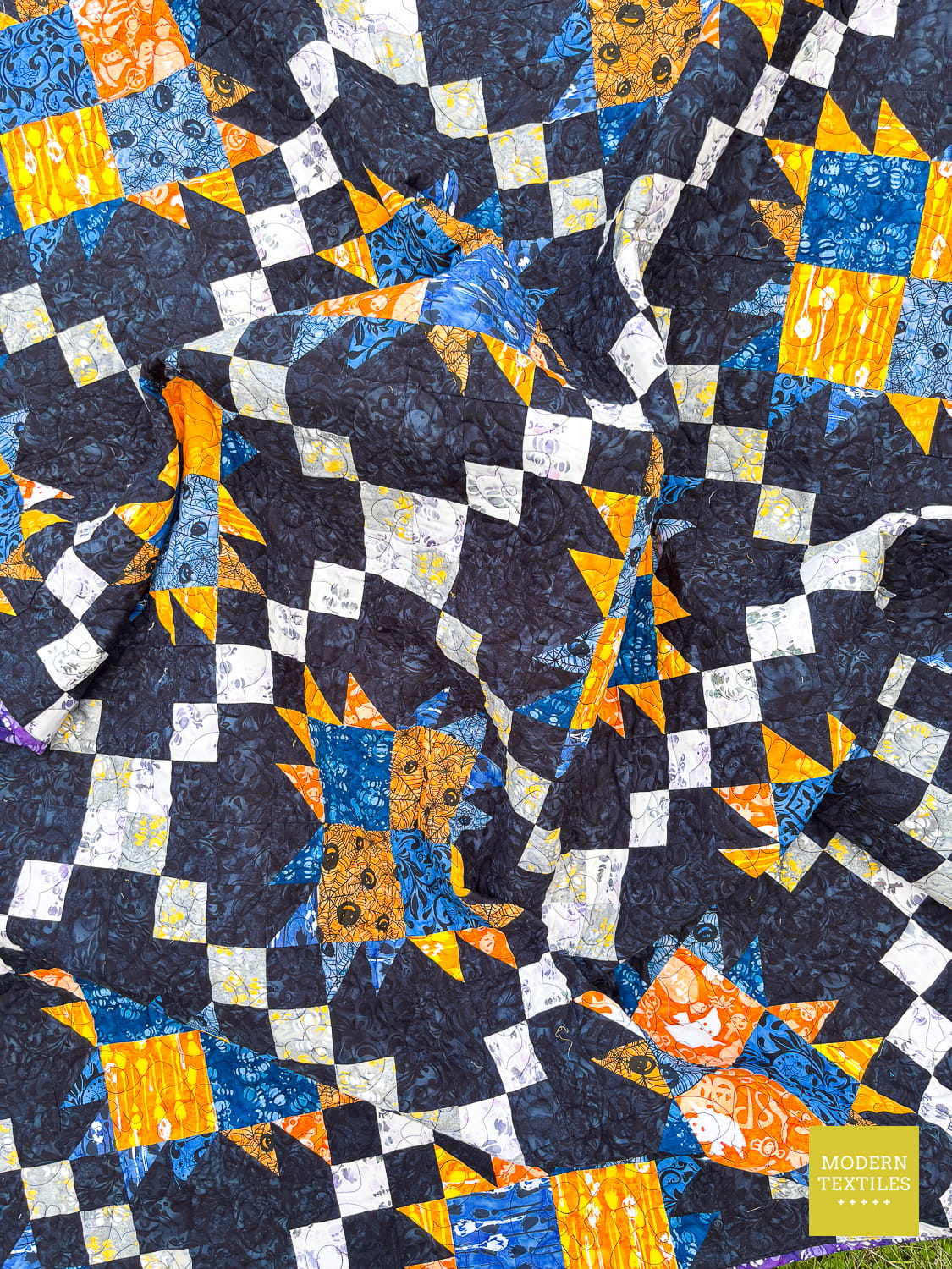 Bear Paw Irish Chain quilt with blue, orange, and white pattern on a black background crinkled to show the teture of the quilt.