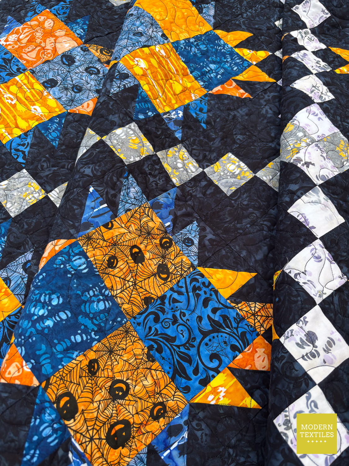 Bear Paw Irish Chain quilt with blue, orange, and white pattern on a black background, quilt is folded onto itself and showcasing the halloween themed batiks fabric.