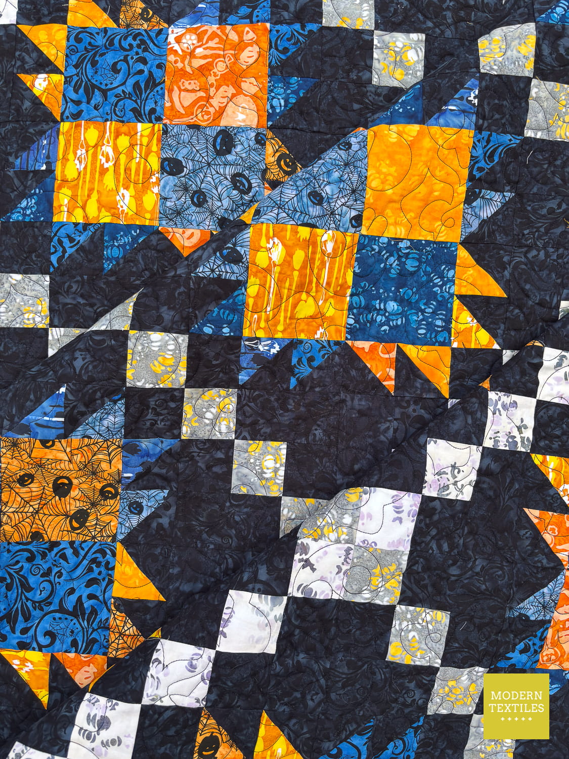 Bear Paw Irish Chain quilt with blue, orange, and white pattern on a black background folded onto itself two times to showcase the drape of the quilt.