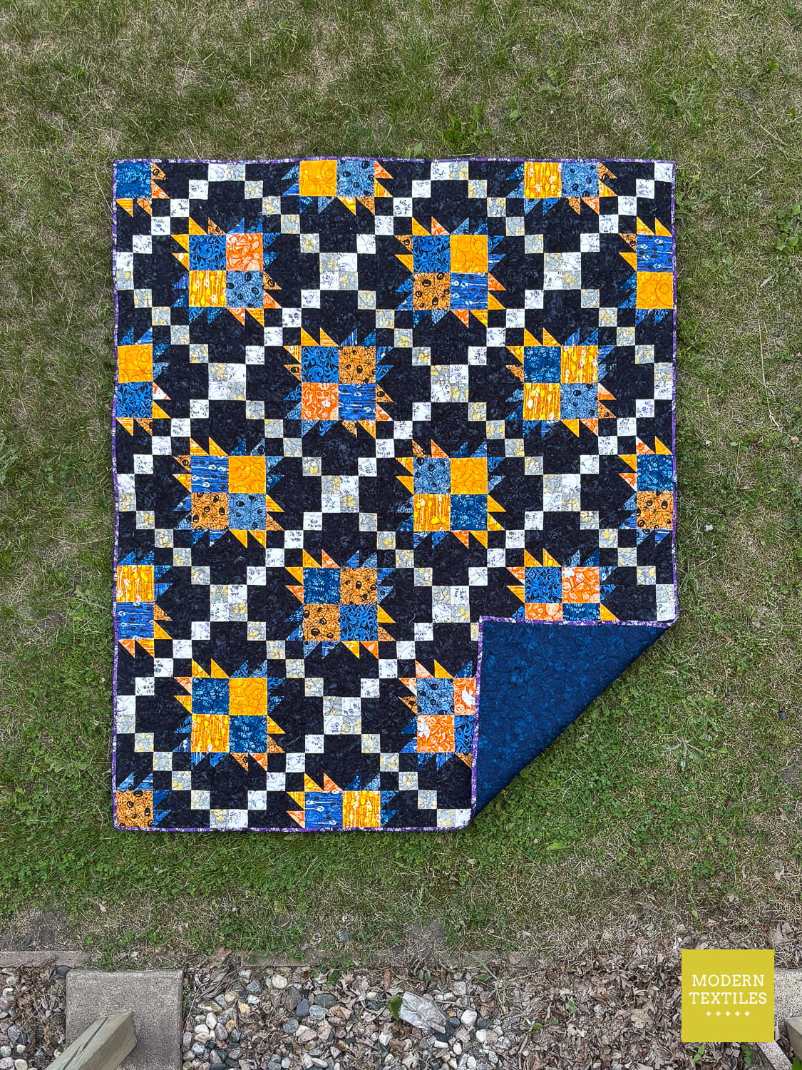 Bear Paw Irish Chain quilt with blue, orange, and white pattern with black background fabric on top of grass, quilt has the lower right portion of the quilt flipped on top of the quilt to showcase the blue batik fabric used as the backing material.