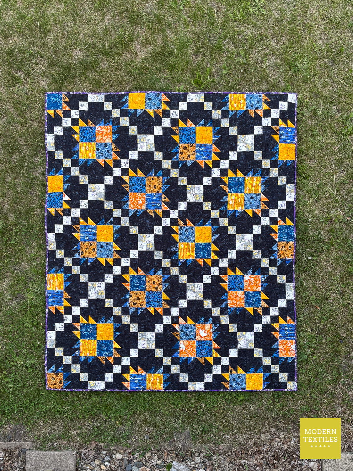 Bear Paw Irish Chain quilt with blue, orange, and white pattern with black background fabric and laid out on top of green grass highlighting the layout of the patchwork blocks of the quilt.