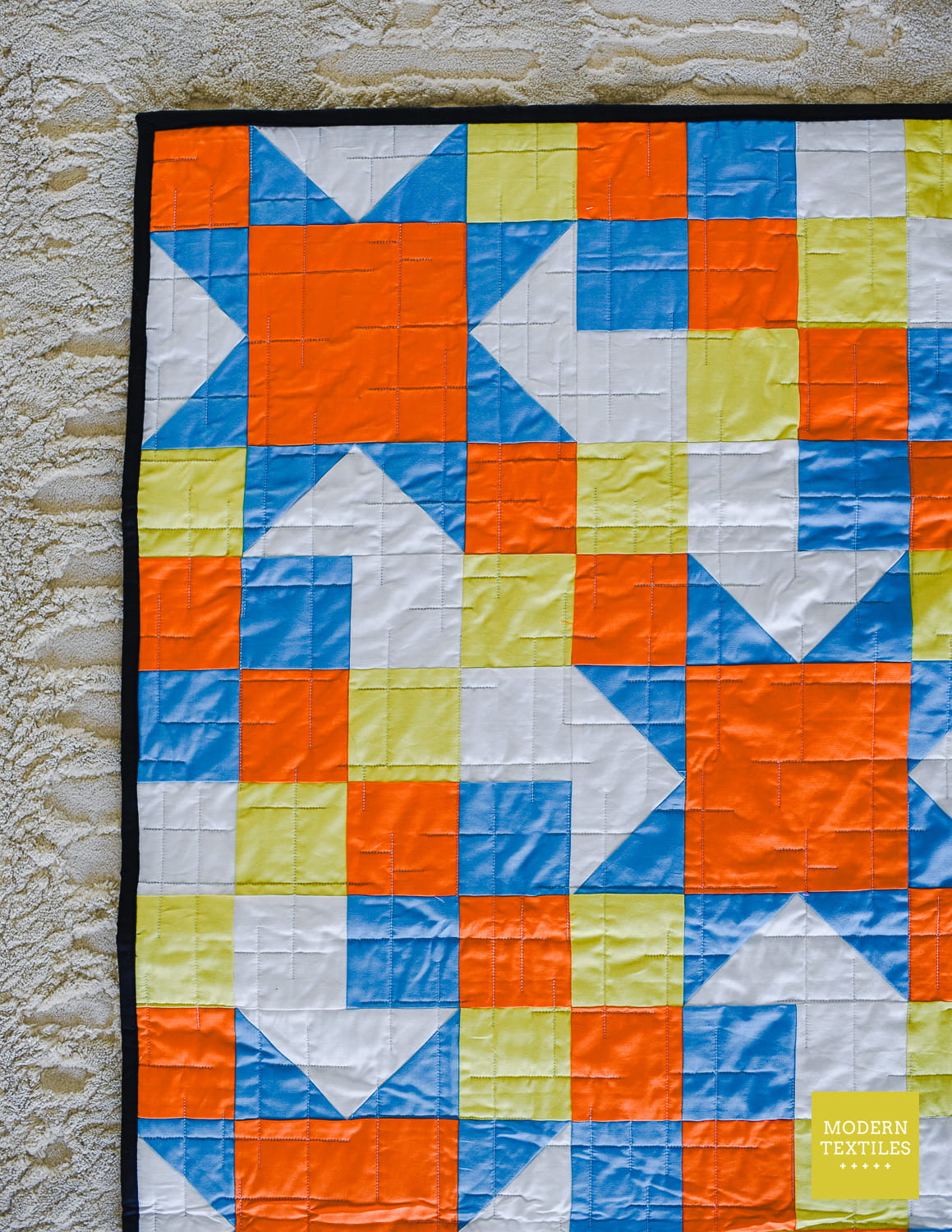 A colorful quilt with a geometric block pattern in orange, blue, white, and pale yellow, shown hanging against a textured beige rug. A small 'Modern Textiles' logo is visible in the bottom right corner.