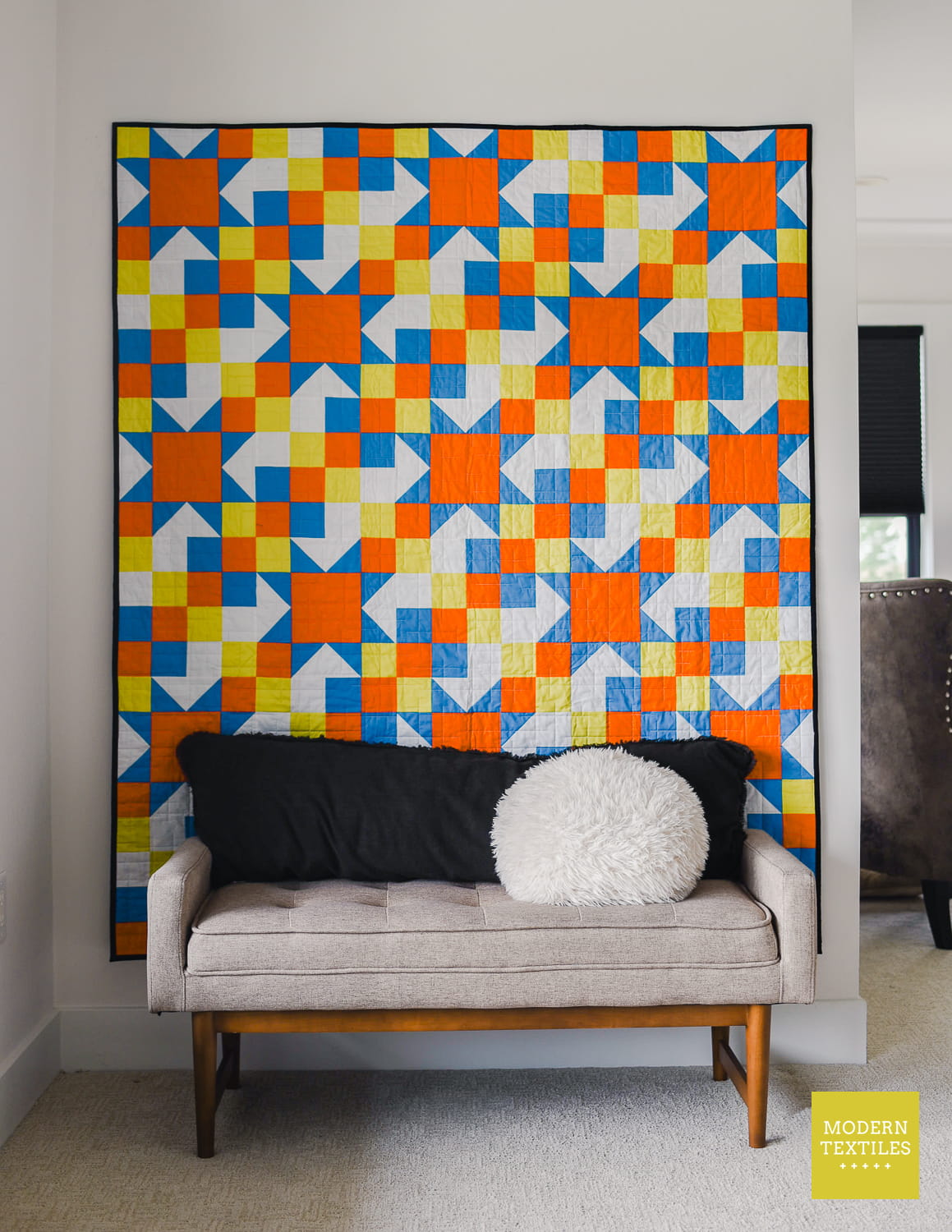 A large quilt hanging with a bold geometric star-block pattern in orange, blue, yellow, and white, displayed above a light beige bench with black and white pillows in a styled interior room.