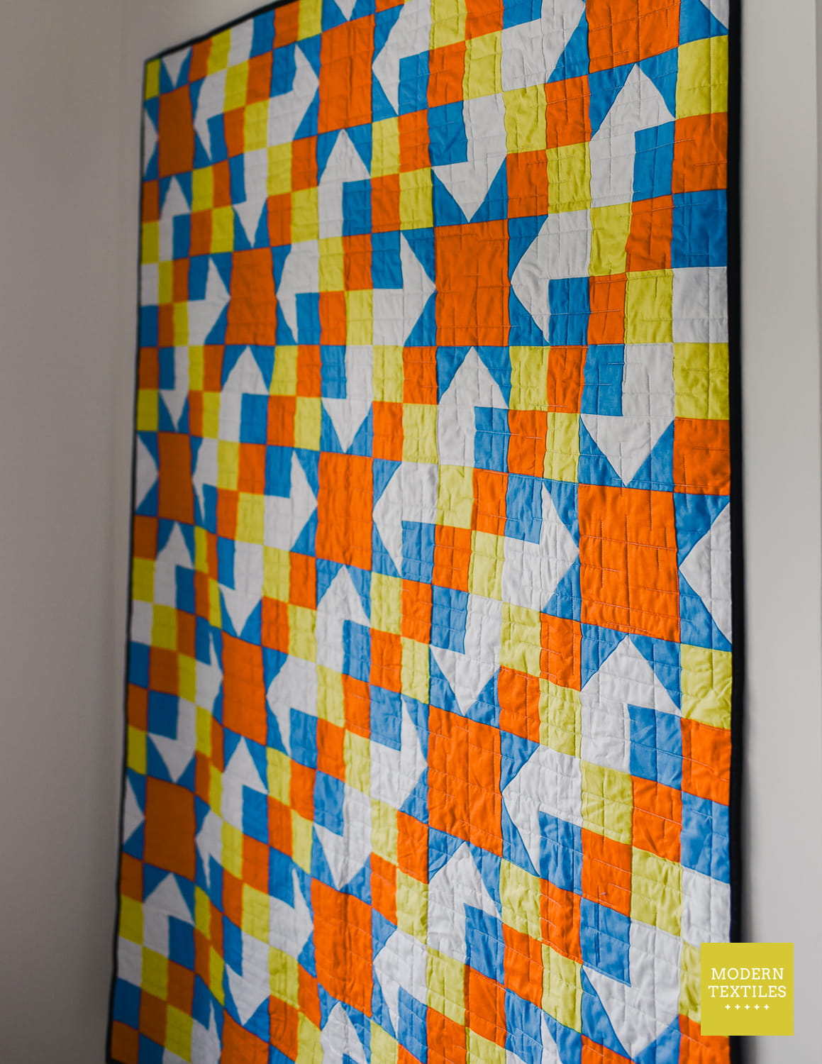 A large geometric quilt hanging on a light gray wall. The quilt features a repeating pattern of orange, blue, yellow, and white stitched fabric blocks.