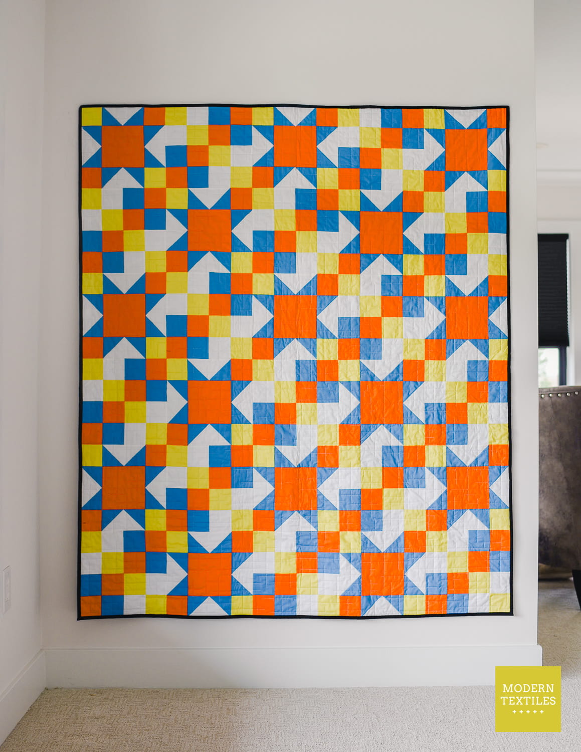 A colorful geometric quilt hanging on a white wall in a room, featuring a repeating pattern of orange, blue, yellow, and white blocks with black borders. A 'MODERN TEXTILES' logo is visible in the bottom right corner.