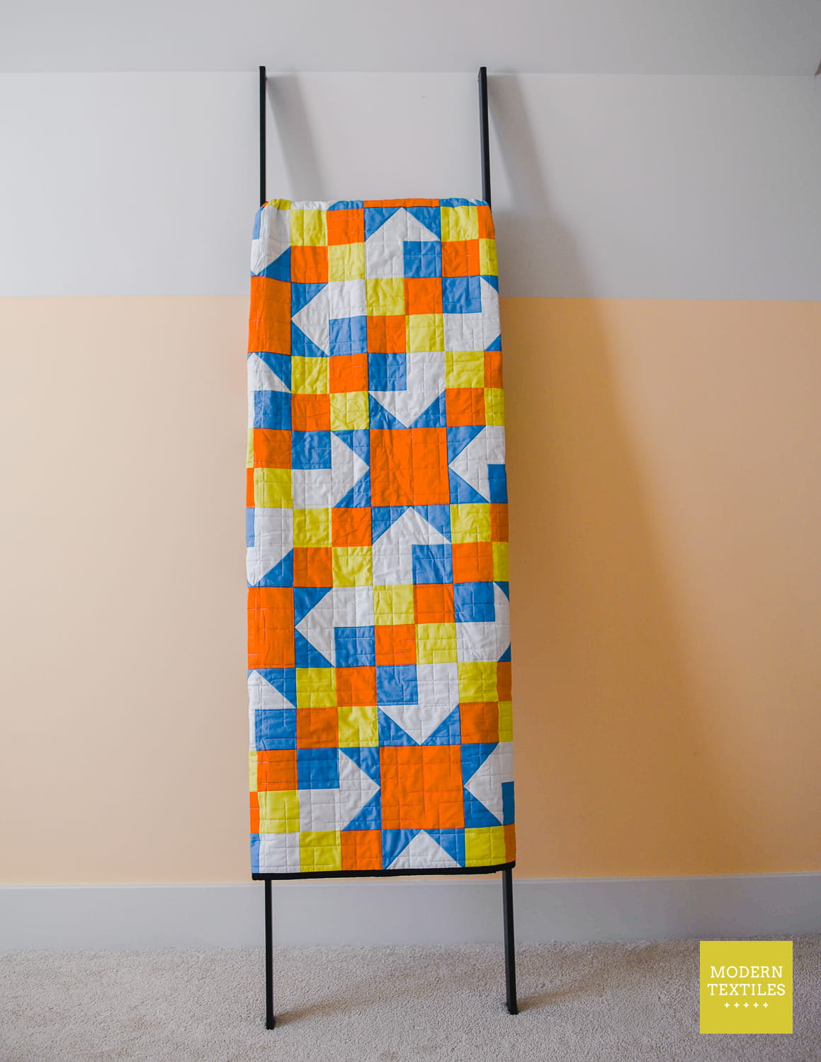A colorful geometric quilt hanging on a black metal ladder stand against a two-tone wall with white and peach sections.