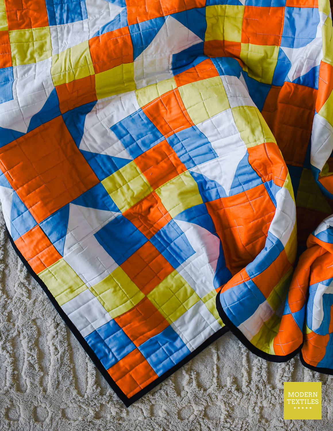 A colorful geometric quilted blanket with a modern patchwork design in blue, orange, yellow, and white, draped over a textured light gray blanket. A yellow 'MODERN TEXTILES' logo is visible in the bottom right corner.