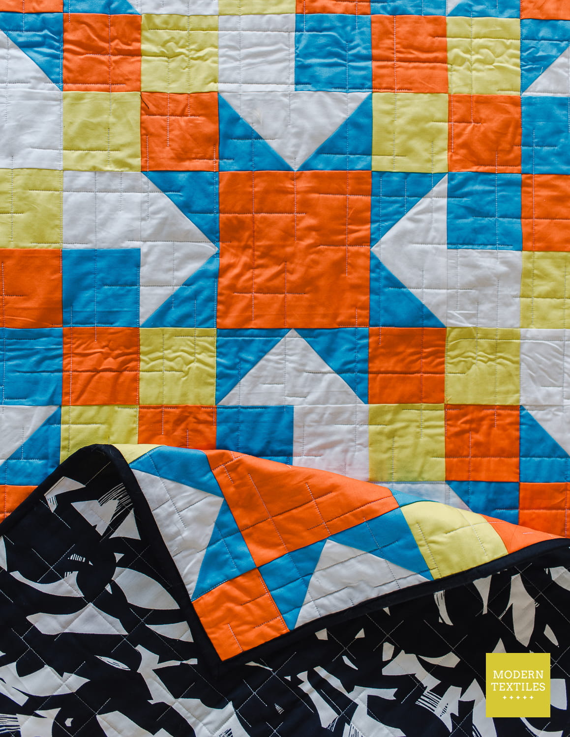 A colorful geometric quilt with orange, blue, white, and pale yellow blocks, shown with one corner folded over to reveal a black and white patterned reverse side. A small yellow 'Modern Textiles' label is visible in the lower right.
