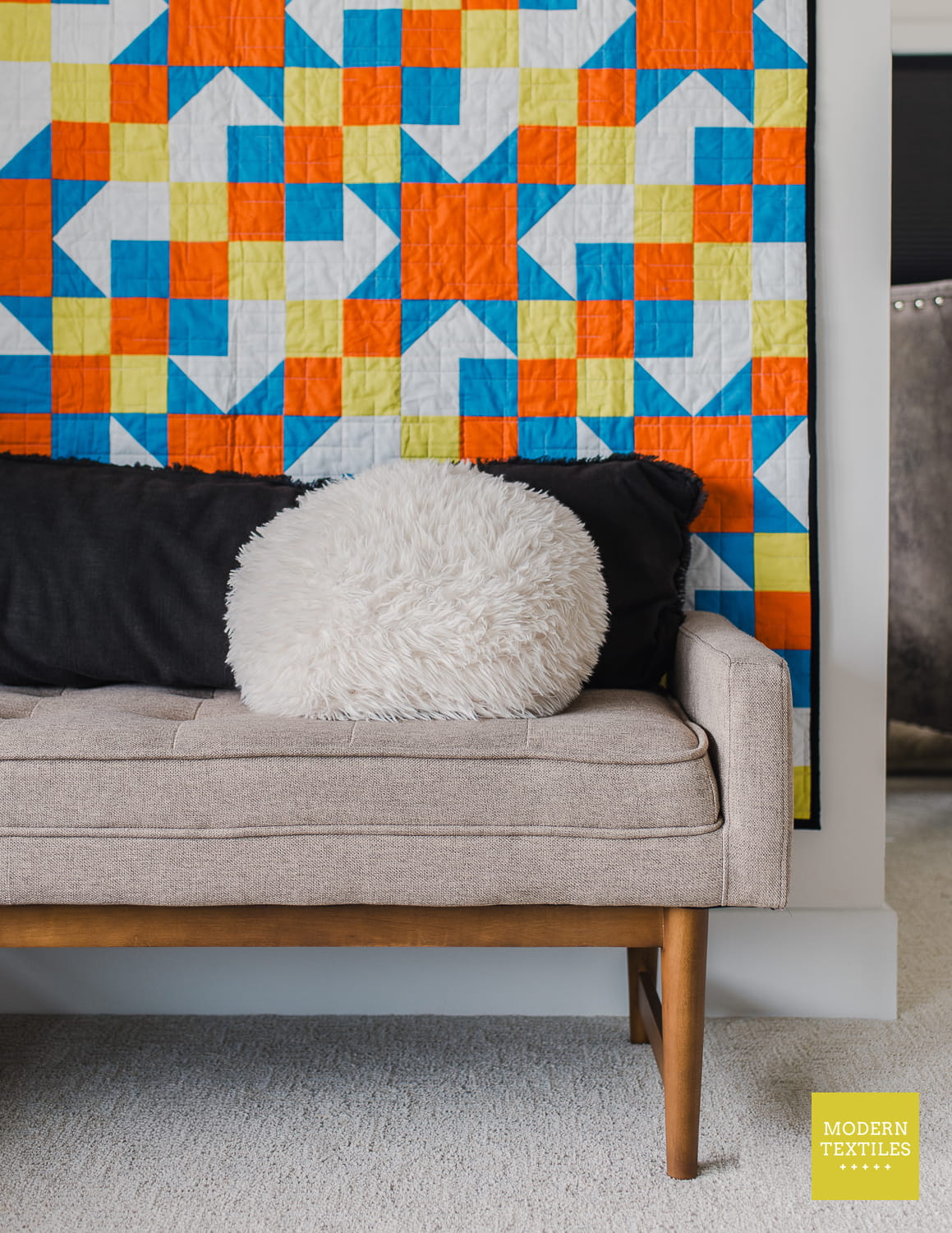 A styled interior scene showing a light beige upholstered bench with wooden legs, decorated with a black pillow, a white shaggy round pillow, and set against a colorful geometric quilt hanging on the wall. A yellow Modern Textiles logo is visible in the bottom right corner.