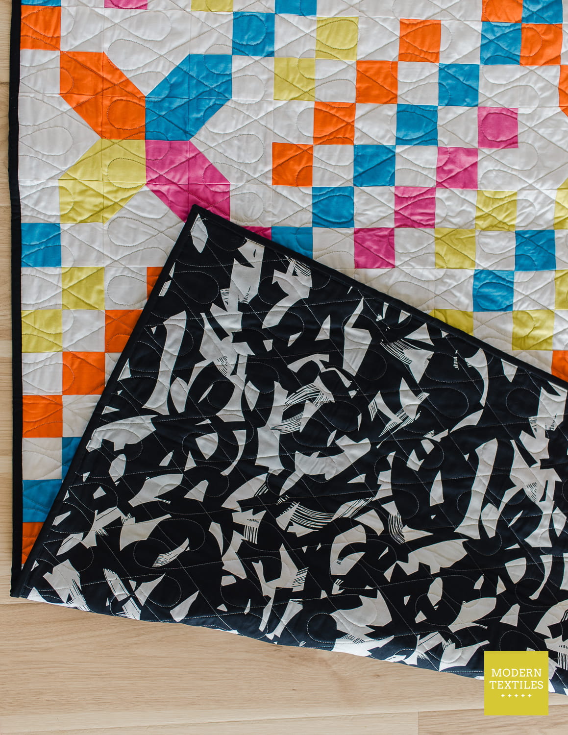 Checkered Crossing quilt laid on light color wood floor, quilt is folded over itself to showcase the brushstrokes backing fabric and black binding against the bright colored quilt. Modern Textiles logo in bottom right corner.