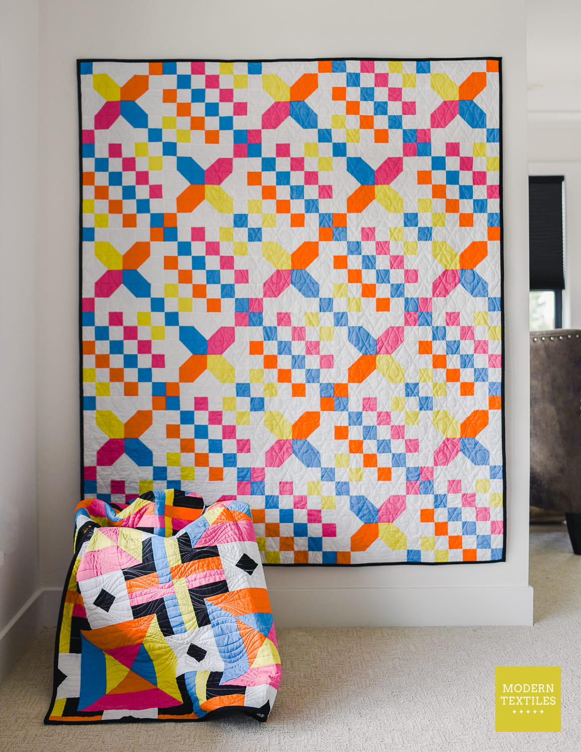 Checkered Crossing quilt hanging on white wall in background. Woven Squares quilt draped in basket in foreground. Modern Textiles logo in bottom right corner.