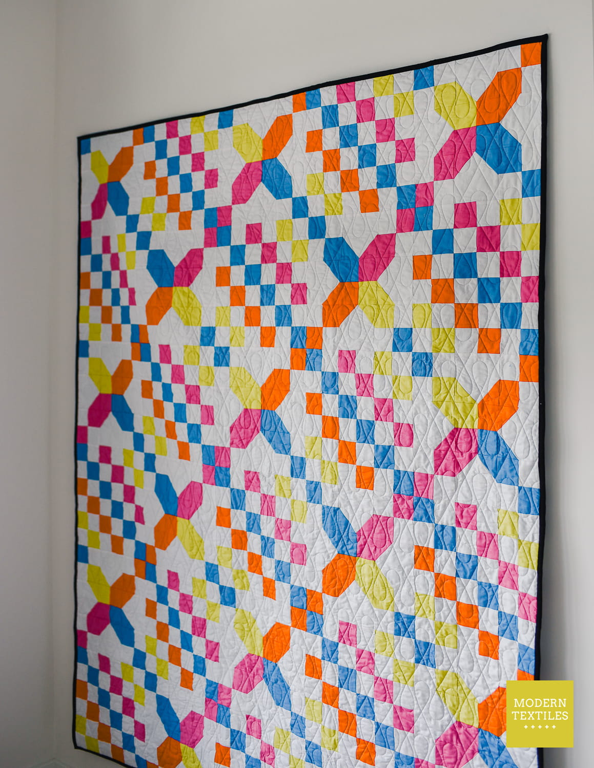 Checkered Crossing quilt hanging on white wall, shown at an angle to highlight the wishbone pantograph quilting texture. Modern Textiles logo in bottom right corner.